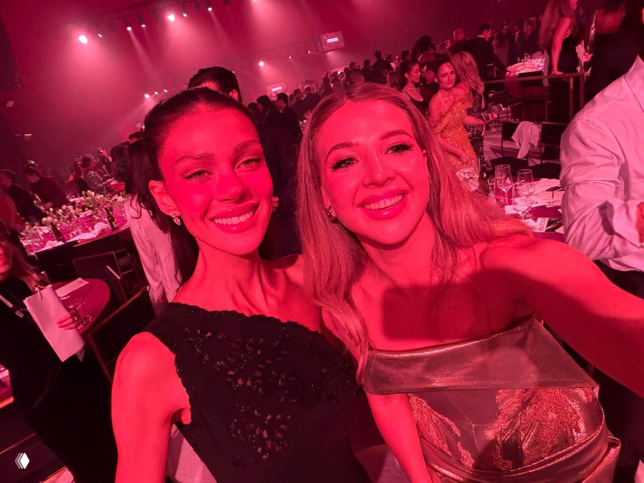 Селфи двух женщин и гостей в красном сценическом освещении на афтерпати; видны столы, бокалы и оживлённая атмосфера