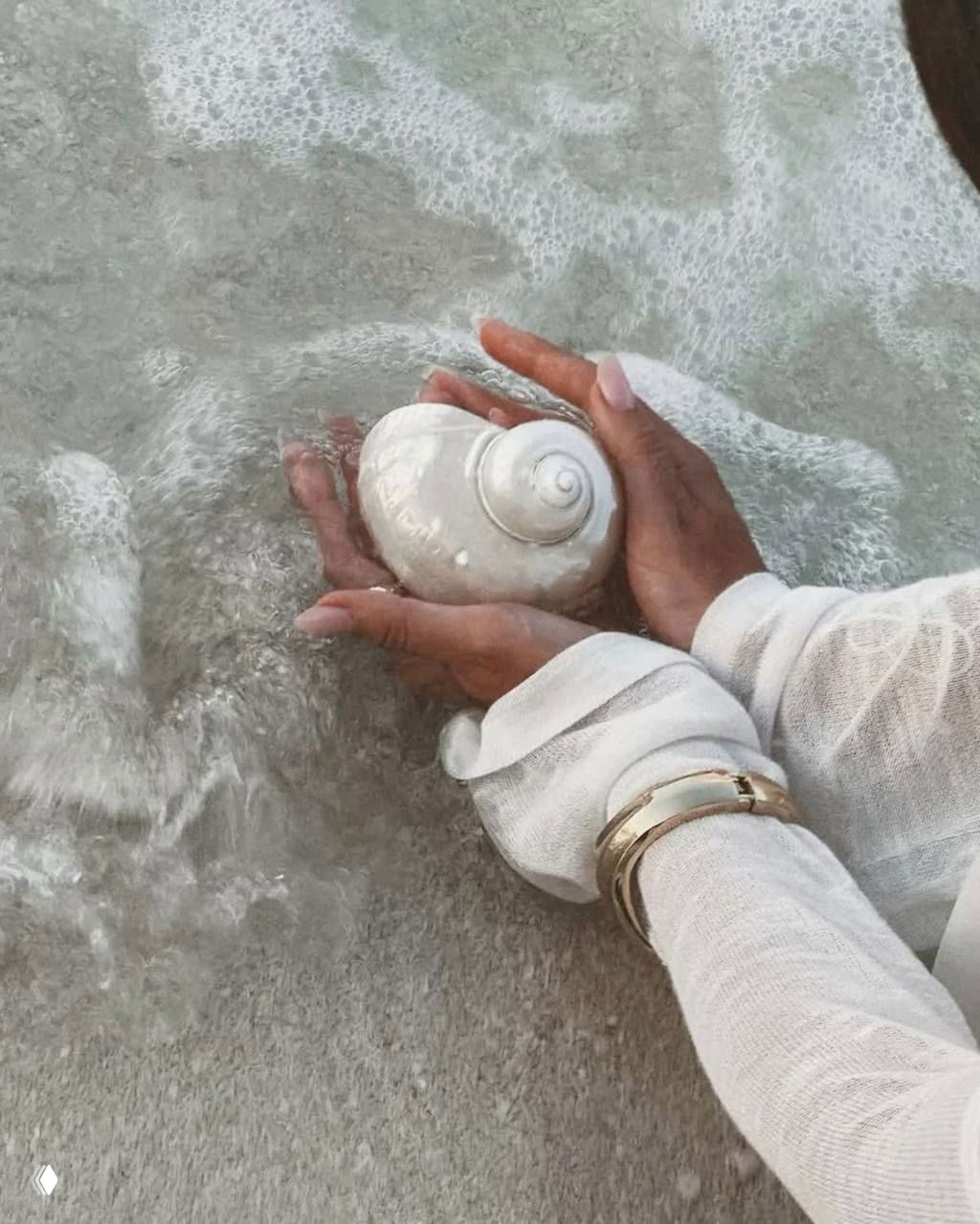 Руки женщины в белой блузе, держащие морскую ракушку на фоне пены и берега — акварельная, нежная цветовая палитра