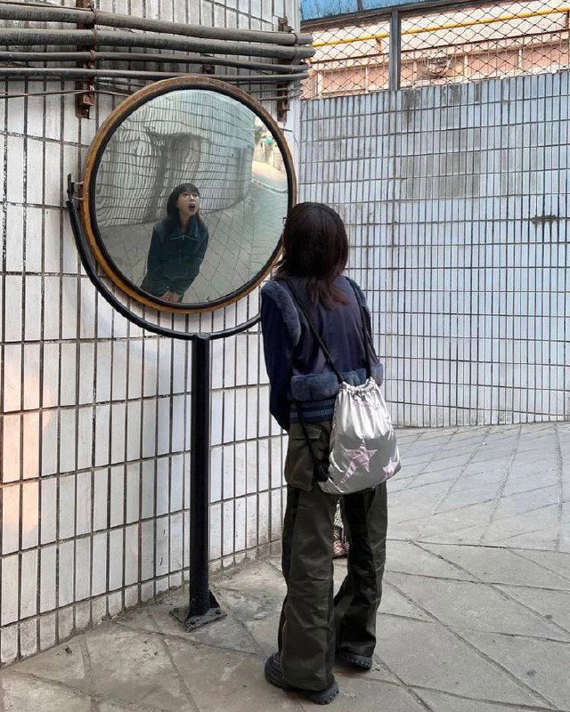 Девушка стоит перед большим выпуклым зеркалом на улице, отражение искажено, кадр предлагает идею странного отражения для фото.