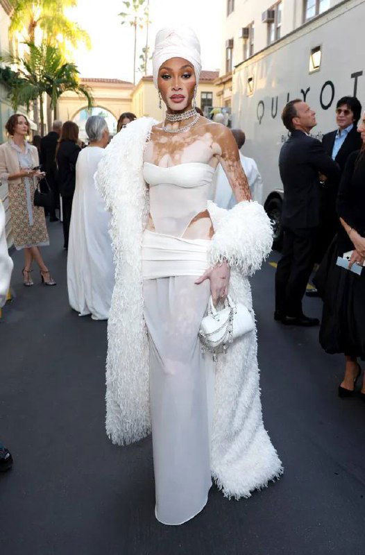 Знаменитость в белом облегающем платье со структурным элементом и пушистым манто позирует на красной дорожке у входа в мероприятие.