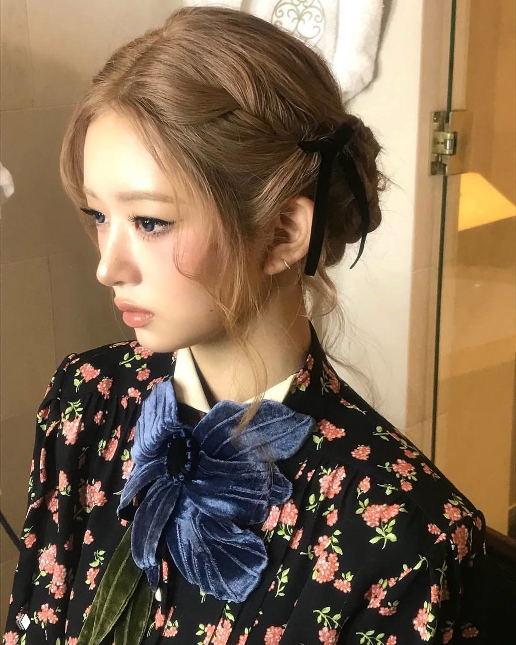 Профиль девушки с аккуратной собранной прической и плетением, украшенной чёрным бантом; деликатный мейкап и цветочный наряд создают вдохновляющий образ.