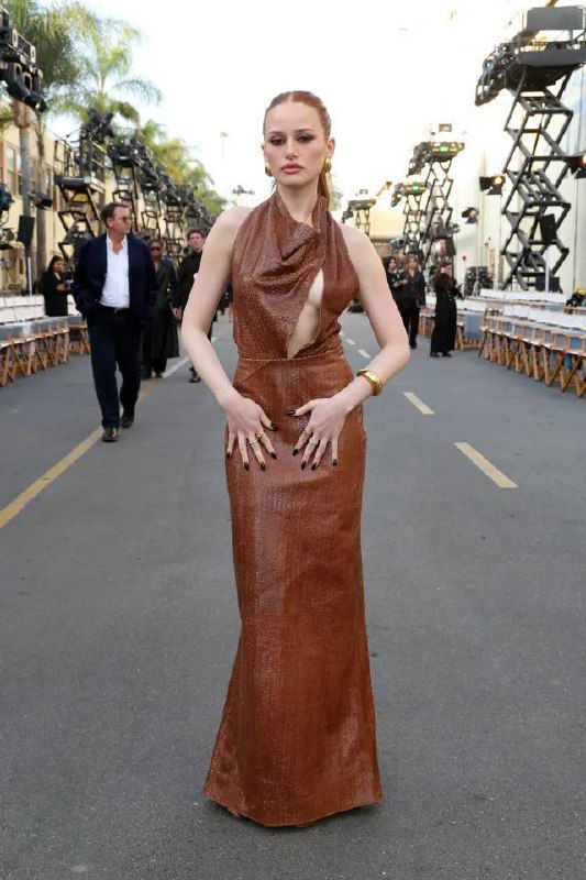 Знаменитость в коричневом длинном платье с асимметричным вырезом стоит на дорожке показа, за ней видно осветительные конструкции и зрителей.