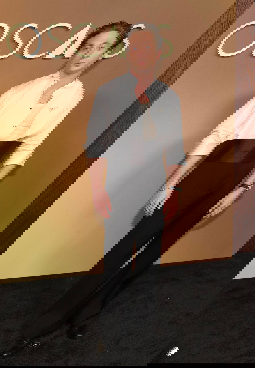 Мужчина в белой рубашке и чёрных брюках с широкой чёрной кушакой на красной дорожке Governors Awards, фото в полный рост на фоне логотипа, фотография с Governors Awards в Лос-Анджелесе.