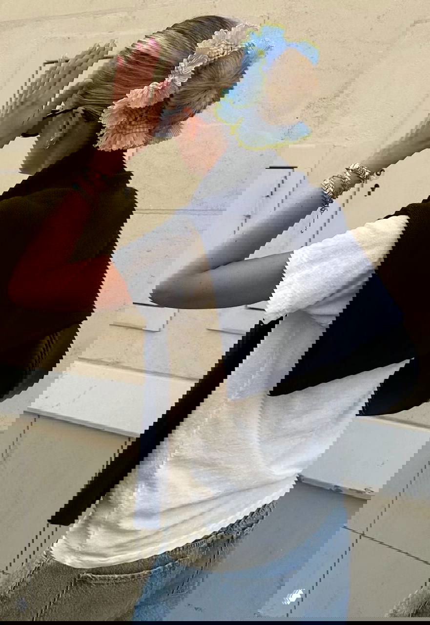 Женщина в повседневном образе с крупной синей цветочной резинкой-скранчи на пучке: уличный фон, акцент на аксессуаре и силуэте прически.