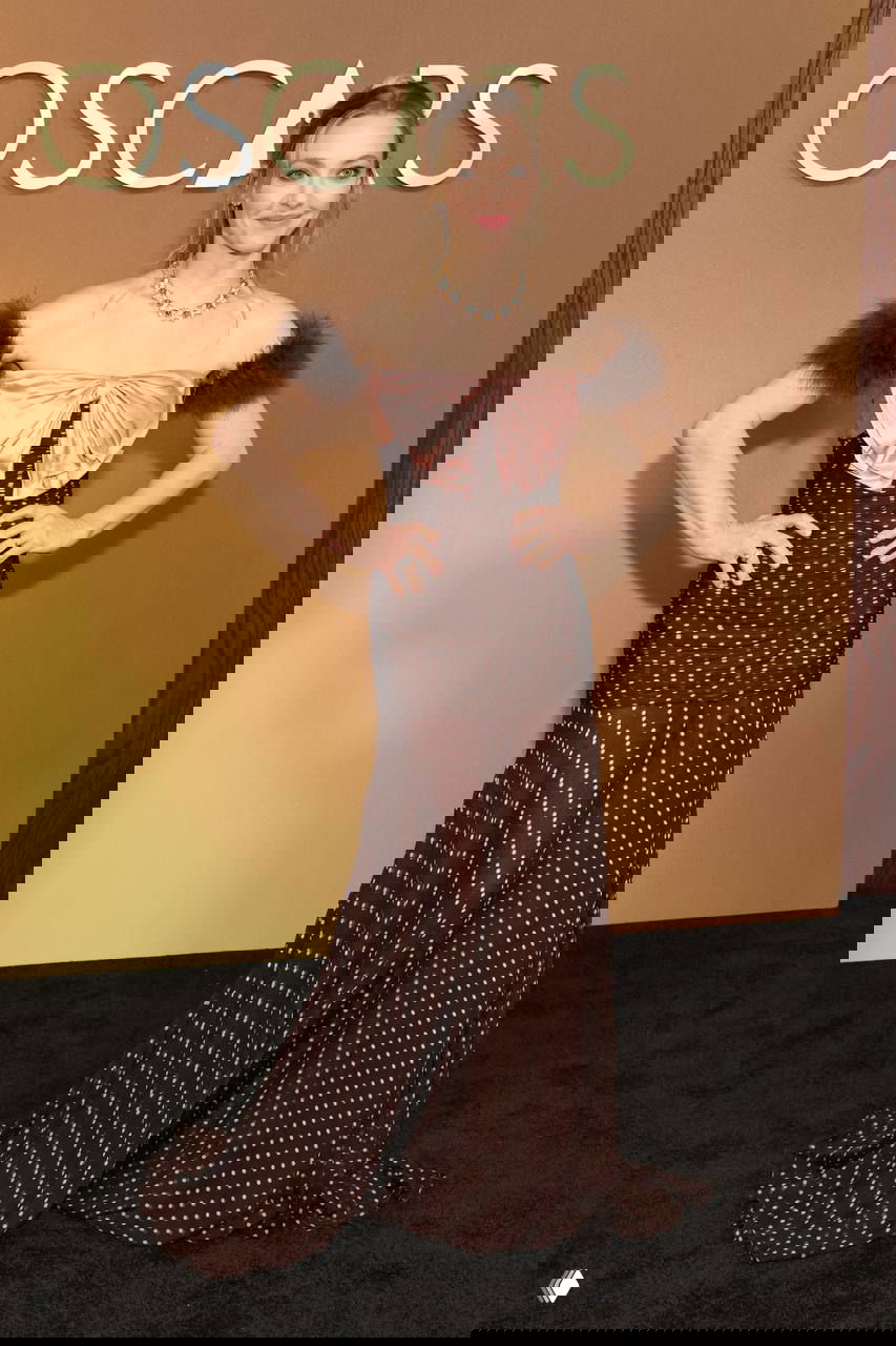 Женщина в коричневом платье в горошек с пушистыми вставками на плечах на красной дорожке Governors Awards, позирует у фона с логотипом, фотография с Governors Awards в Лос-Анджелесе.