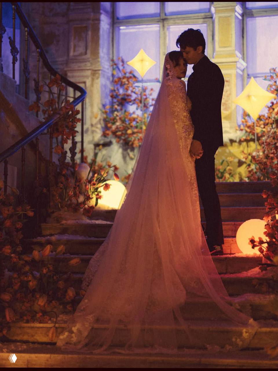 Пара стоит на лестнице: невеста в длинной фате и жених в тёмном костюме, тёплая подсветка и цветочные композиции создают романтическую атмосферу.