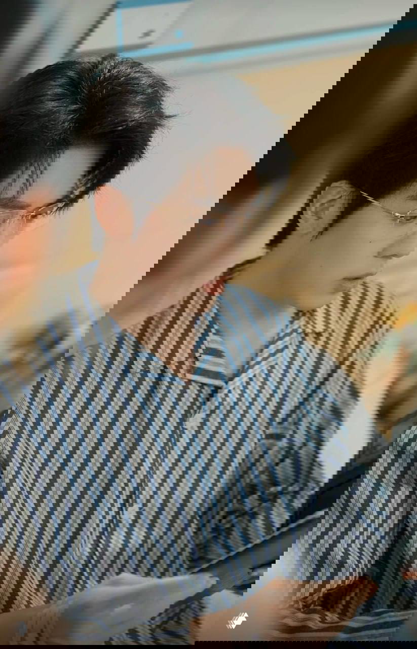 Кадр с молодым мужчиной в полосатой пижаме и очках, он рассматривает планшет или книгу, сцена в палате или комнате — спокойный бытовой эпизод.