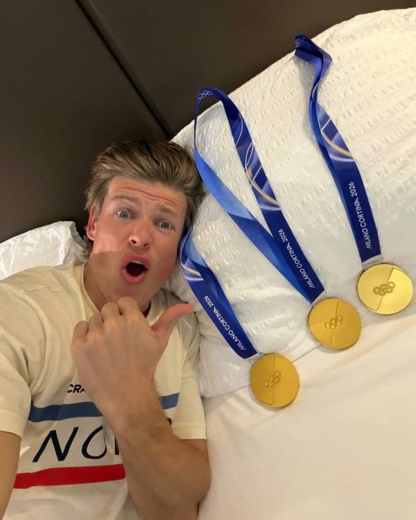 Радостный спортсмен демонстрирует олимпийские медали на кровати: крупный план эмоций и золотых наград, фотокадр с реквизитом соревнований.