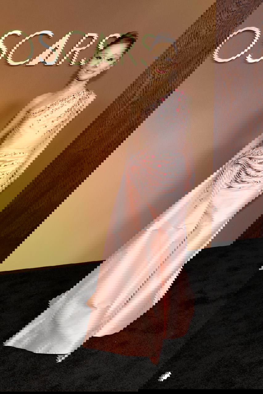 Женщина в розовом асимметричном вечернем платье на красной дорожке Governors Awards, элегантная поза и украшения на фоне логотипа, фотография с Governors Awards в Лос-Анджелесе.