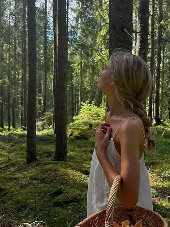 Девушка в лесу стоит среди высоких сосен и мха с плетёной корзиной, погружённая в природную атмосферу и лёгкую невесомость образа.