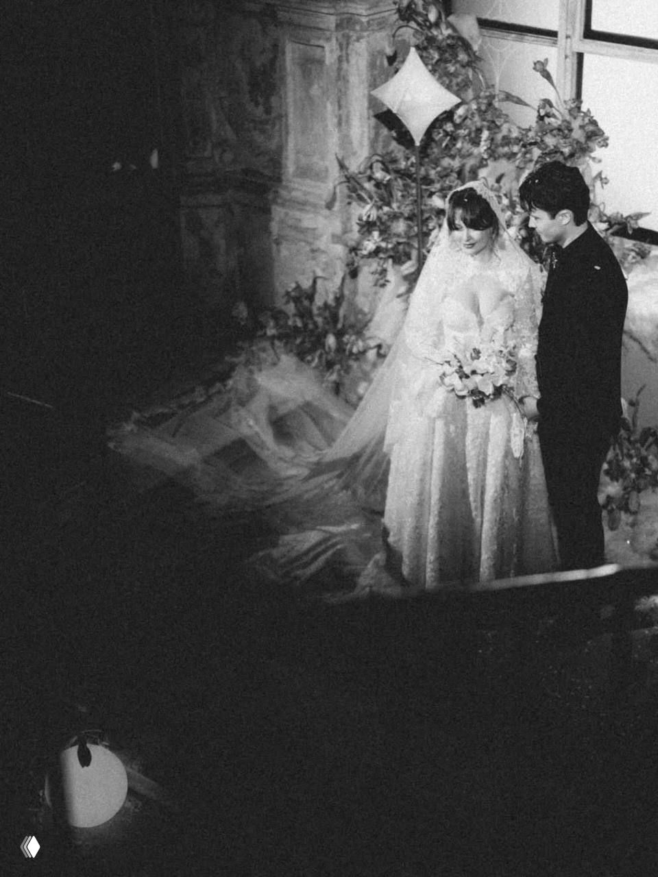 Зернистый чёрно‑белый снимок пары в интерьерном пространстве с цветочными композициями на ступенях, кадр передаёт ретро‑настроение вечера.