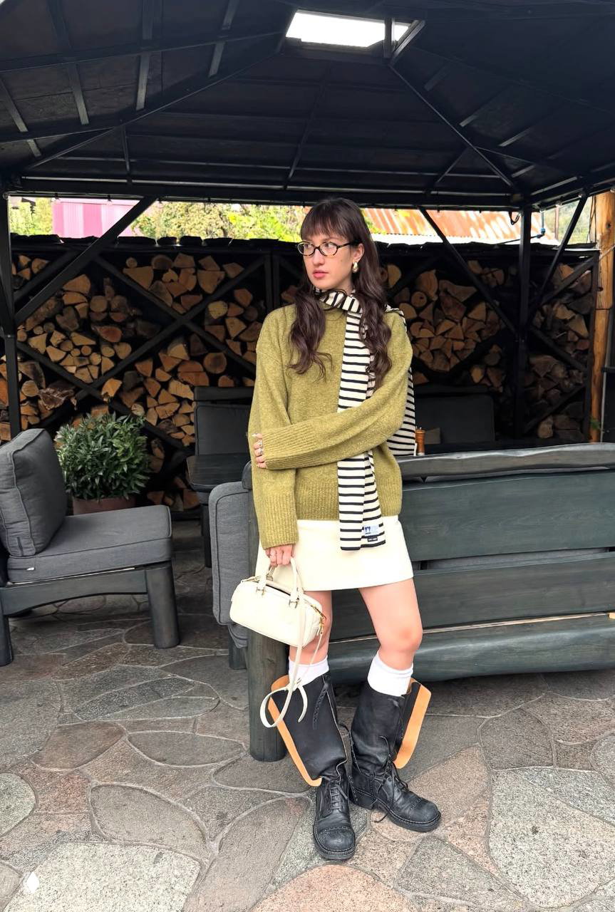 Лиза в оливковом свитере и полосатом шарфе, мини-юбка и грубые ботинки на фоне стога дров и уличной веранды — уютный осенний лук.