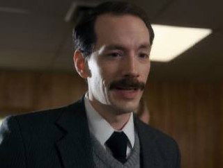 Крупный план мужчины с усами и деловым костюмом в кабинетном интерьере — выразительный фрагмент персонажа из телесериала.