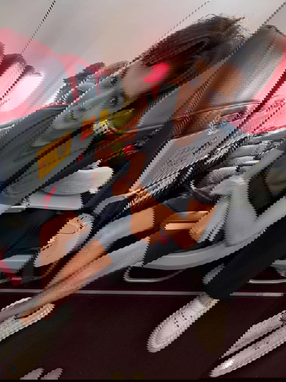 Акмаль сидит в кресле эконом‑класса самолёта, рядом другие пассажиры; видно интерьер салона и красные кресла.