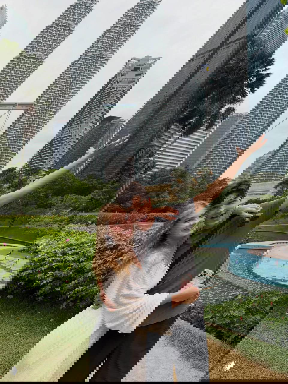 Пара обнимается в парке на фоне башен Петронас в Куала‑Лумпуре: зелёный газон, бассейн и небоскрёбы за ними.