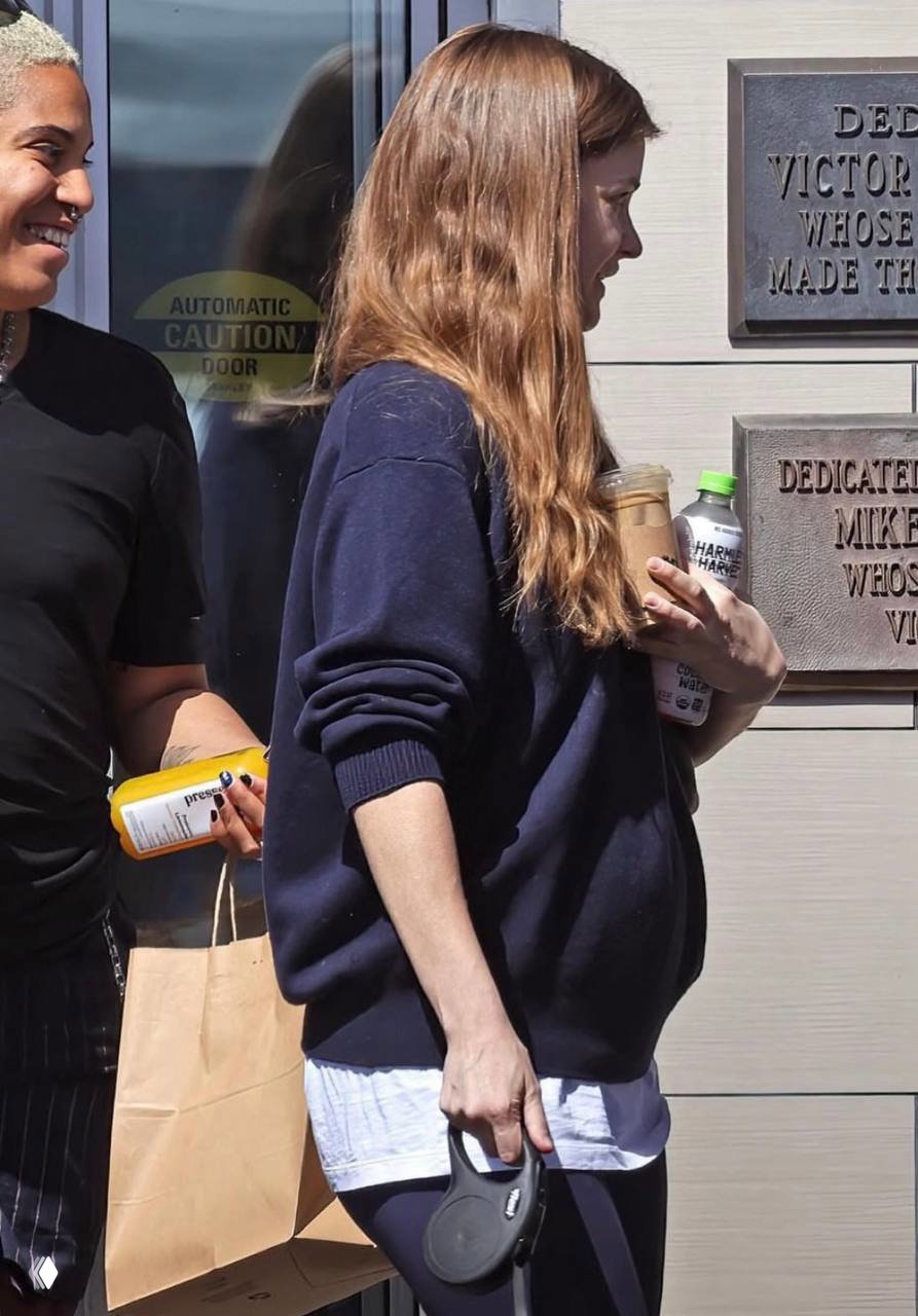 Крупный план Барбары Спроус в профиль: модель держит бутылку и стакан, видно округление живота, кадр сделан во время выхода из кафе/лавки на улице.