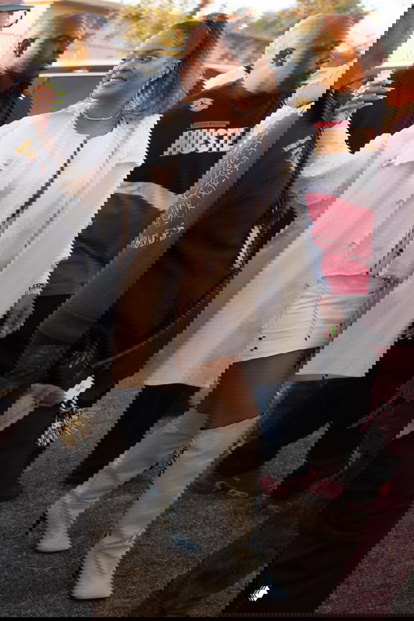 Женщина в короткой белой мини-юбке, удлинённой рубашке и высоких белых сапогах идёт по плотной толпе с охраной, уличная сцена на мероприятии.