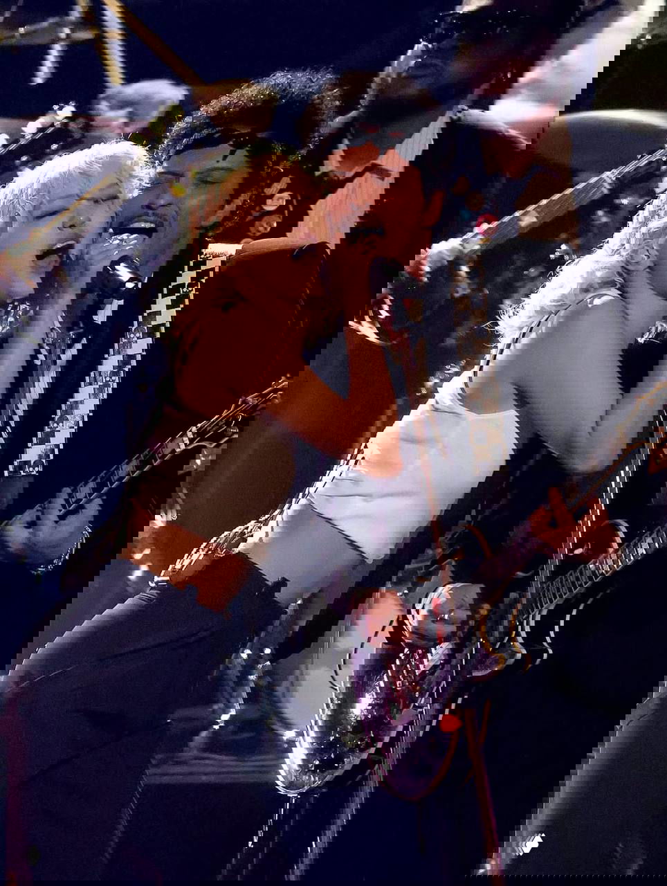 Дуэт музыкантов на сцене: вокалистка и гитарист поют в микрофон на тёмном фоне с концертным освещением и бэк‑группой.