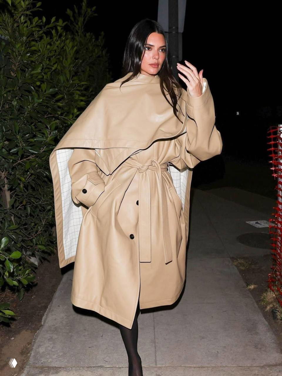 Фигура в бежевом тренче с поясом и широкими рукавами идёт по тротуару рядом с зелёными кустами, городской стритстайл, аксессуары в руках.