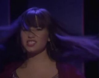 Кадр из фильма «Camp Rock: Музыкальные каникулы»: молодая певица в движении, развевающиеся волосы и эмоциональное выражение лица на тёмном фоне