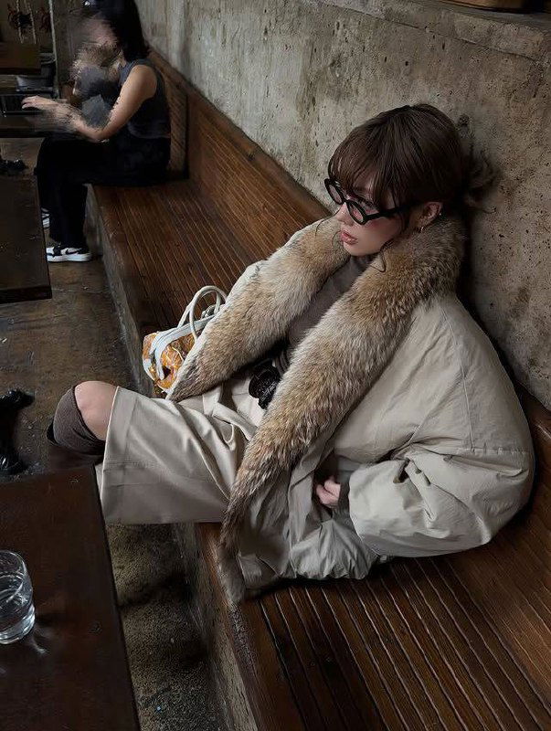 Городской кадр: девушка сидит в бежевой парке с длинной меховой опушкой на воротнике, интерьер кафе подчёркивает повседневный характер образа.