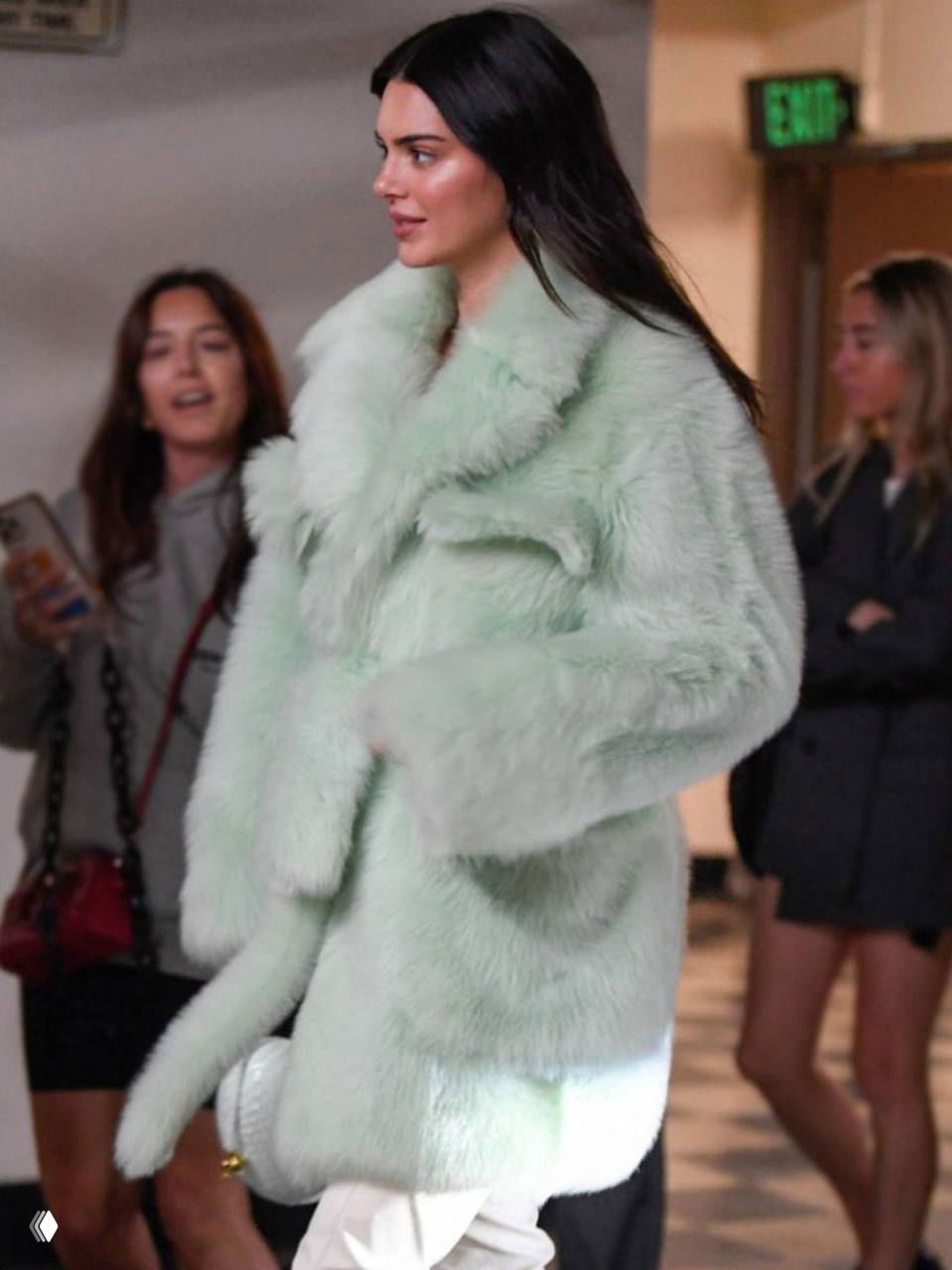 Женщина в мятной пушистой шубе идёт по коридору на светлом полу, прохожие и интерьер вокруг создают контекст уличной моды и тренда.