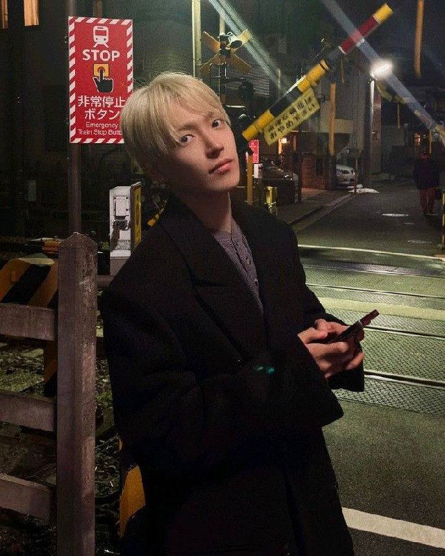 Портрет Хонджуна с осветлёнными волосами на ночной улице рядом с железнодорожным переездом и знаком "STOP", он держит телефон в руках.