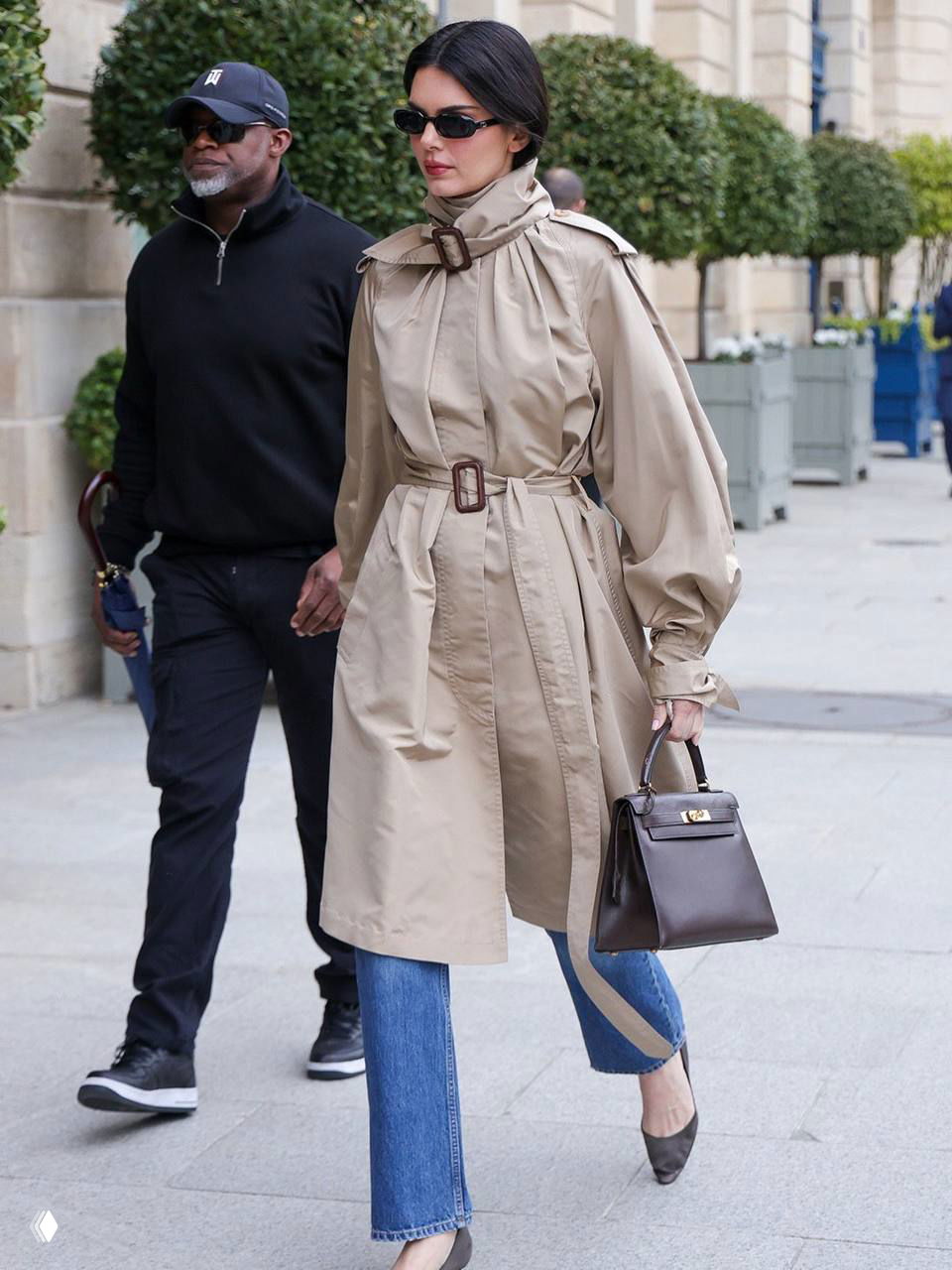 Образ с тренчем, джинсами и темными очками: женщина шагает по городскому тротуару, рядом охрана и прохожие, сдержанный минимализм.