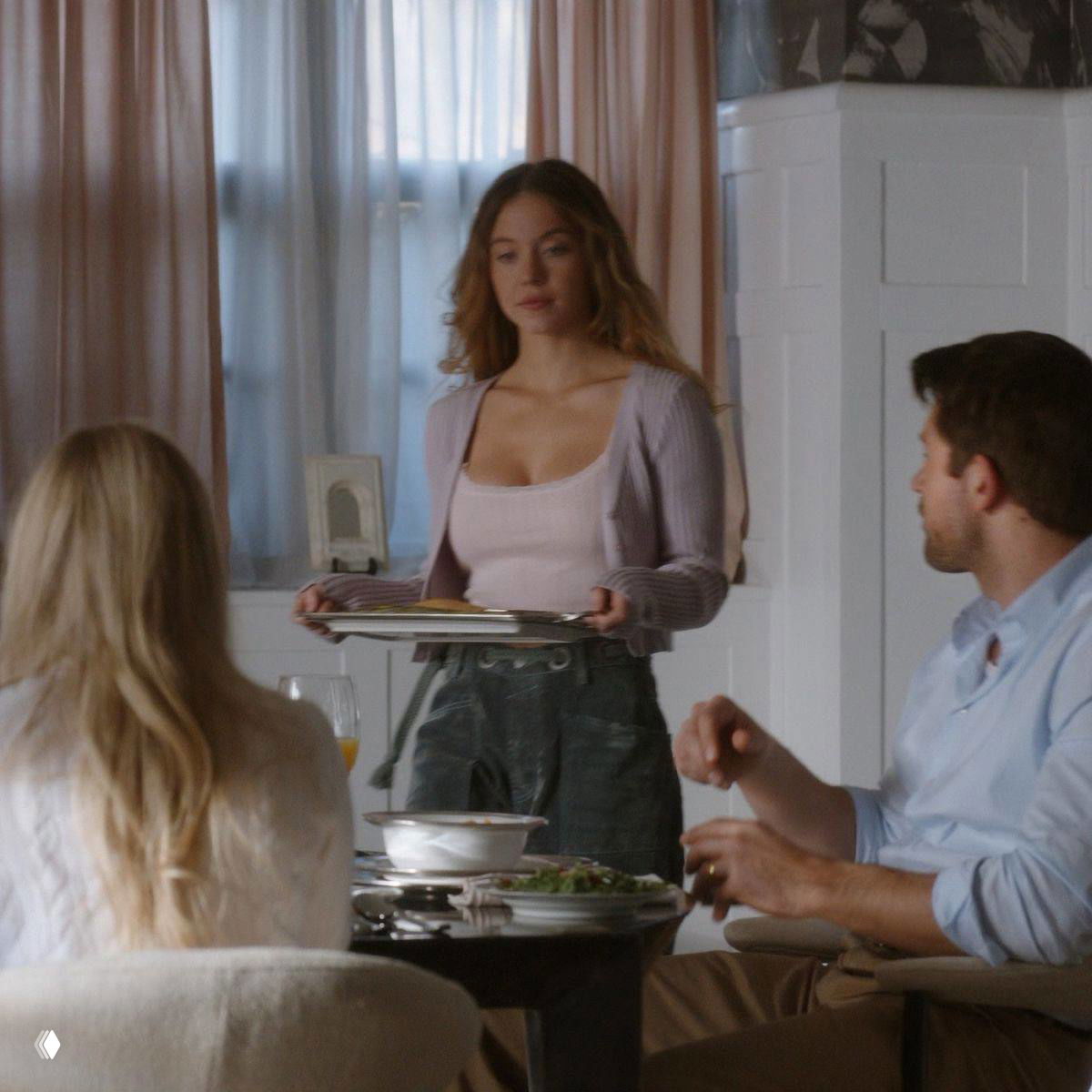 Кадр из фильма: девушка в форме горничной несёт поднос за столом в семейной обстановке; сцена демонстрирует бытовую динамику между героями.