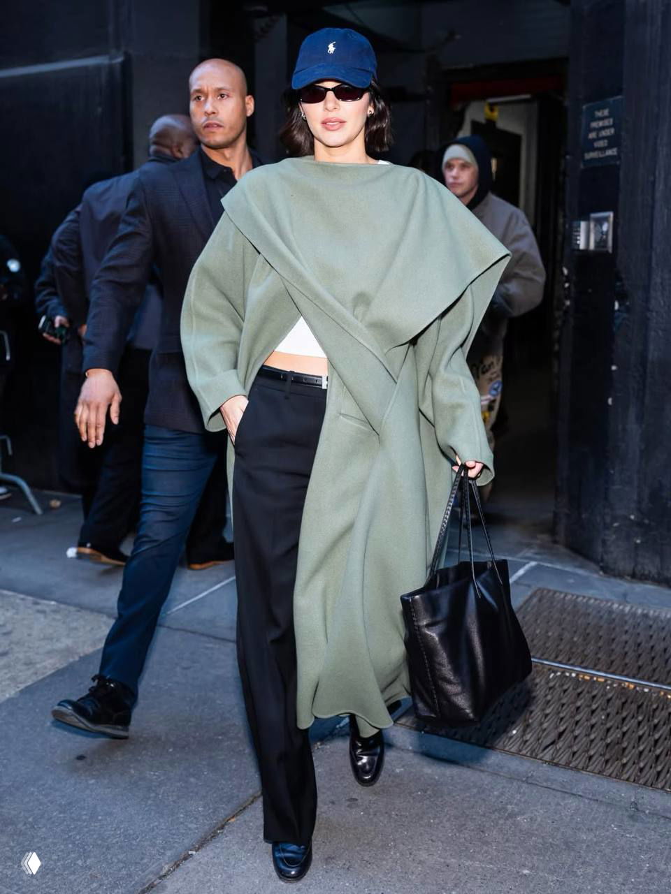 Образ в длинном пальто-пончо светло-зелёного оттенка с чёрной сумкой и кепкой, уличная сцена с охраной и другими прохожими на заднем плане.