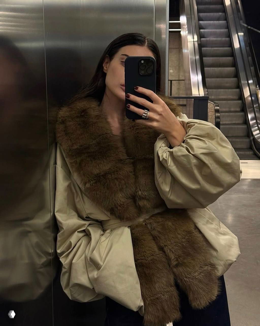 Зеркальное селфи: женщина у эскалатора в куртке с широкой коричневой меховой вставкой, кадр подчёркивает текстуру меха и городской фон.