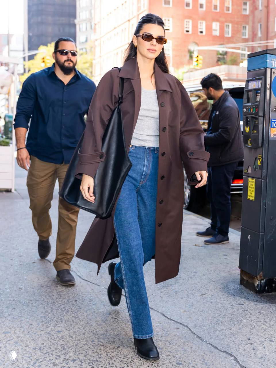 Повседневный городской образ: длинное коричневое пальто, джинсы и большая сумка, девушка идёт по улице в дневной свет, уличные детали вокруг.