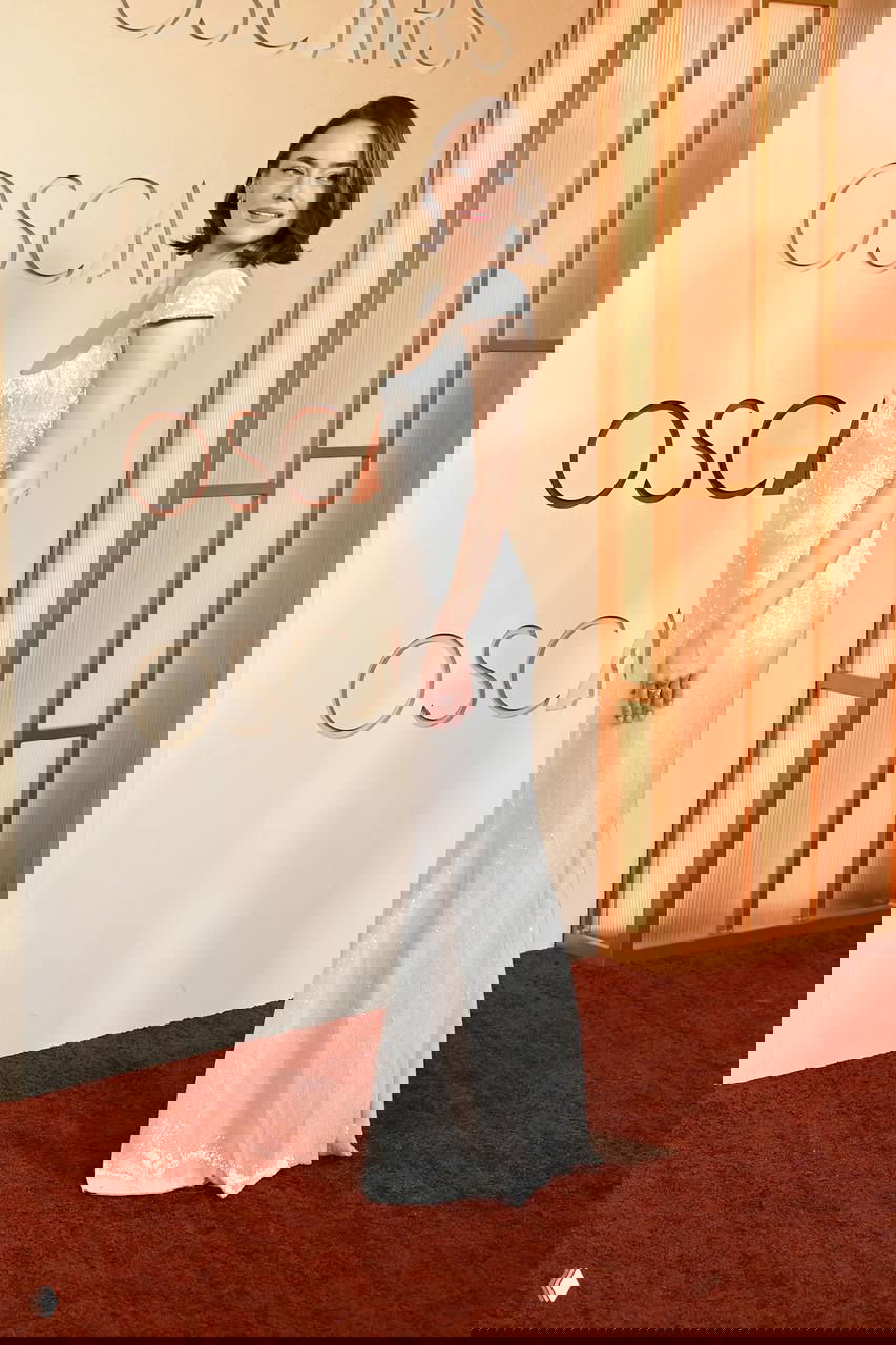 Женщина в узком сверкающем серебристом платье стоит у декора «Оскар», подчёркивая минималистичный элегантный образ на красной дорожке.