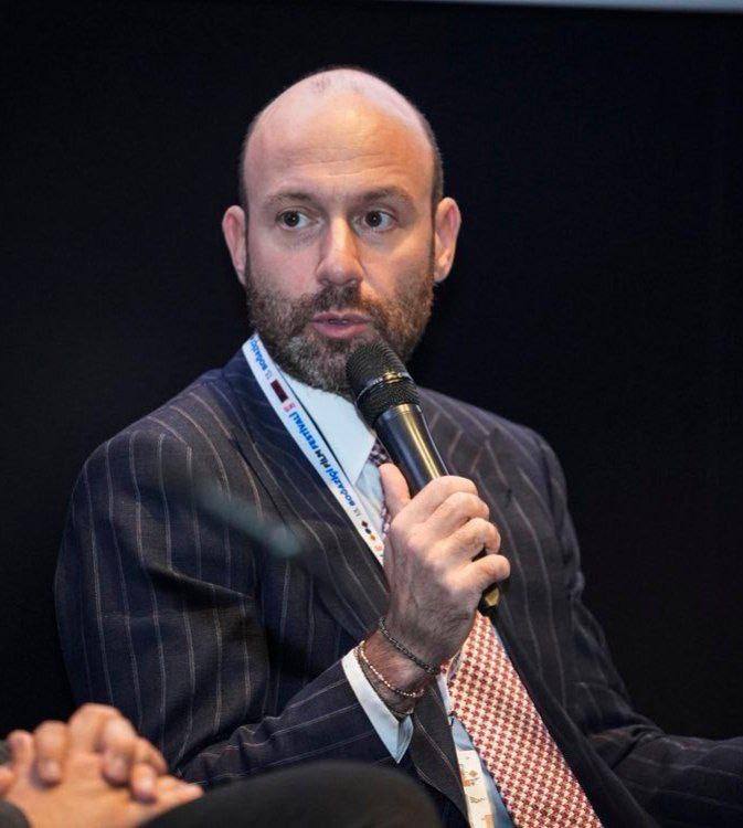 Мужчина в деловом костюме держит микрофон на сцене во время панели или интервью, кадр сделан крупным планом на фоне тёмного зала.