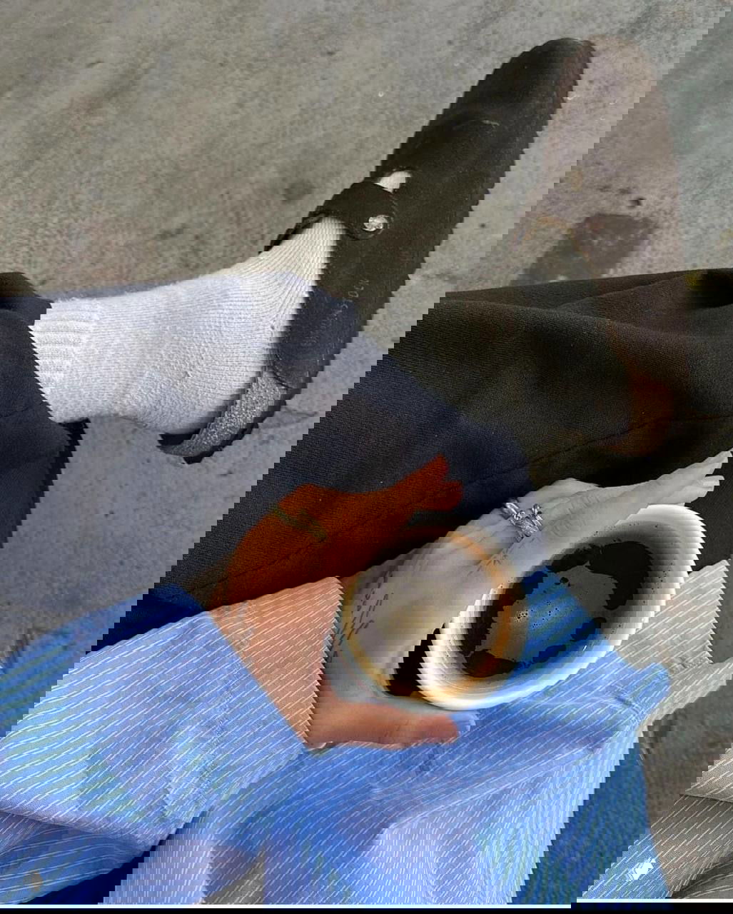 Крупный план: нога в светлых носках и тёмных замшевых биркенштоках, в руке бумажный стаканчик с чёрным кофе — уютная городская сцена.