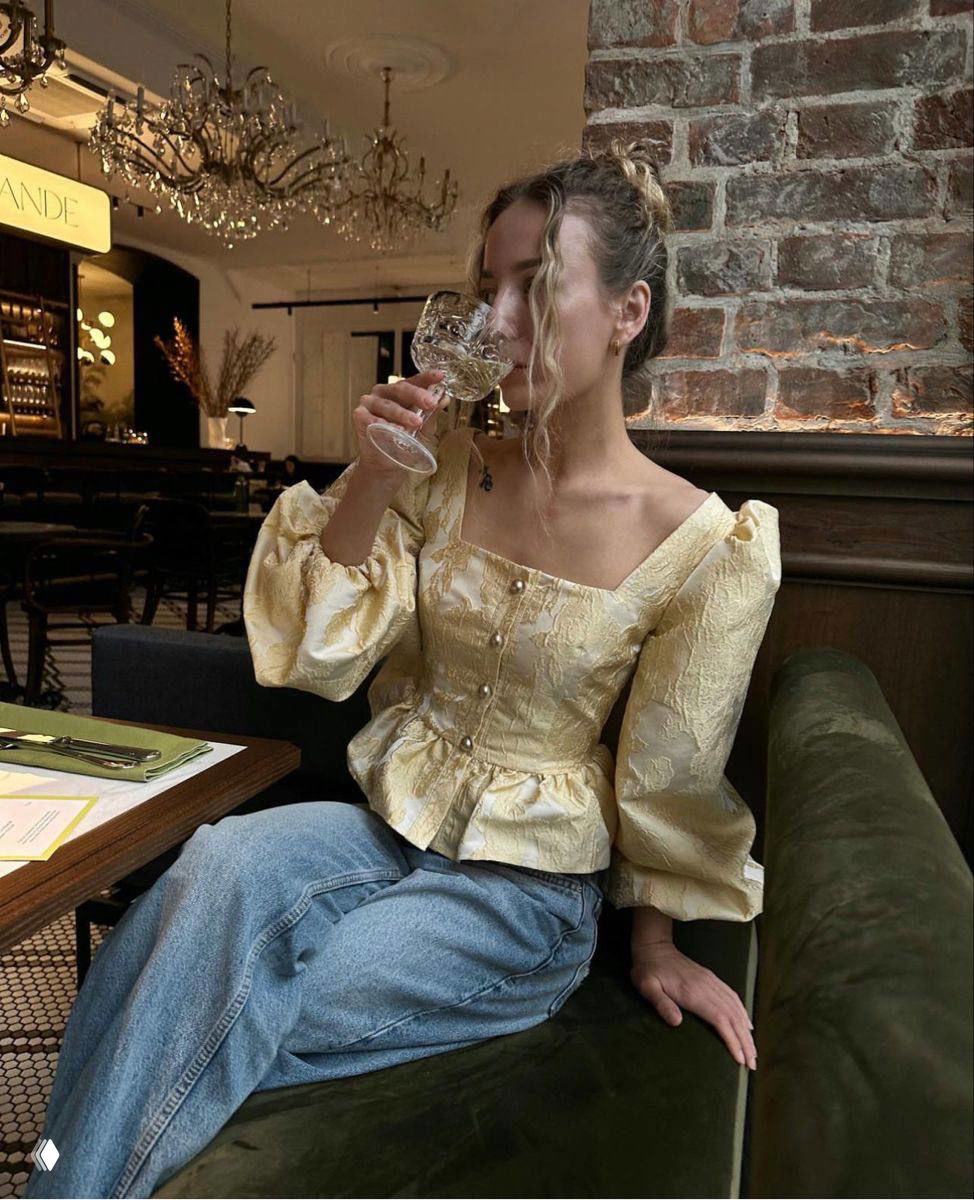 Женщина в ресторане в кремово‑жёлтой жаккардовой блузе с пышными рукавами и жемчужными пуговицами, сочетание винтажного кроя и современного образа.