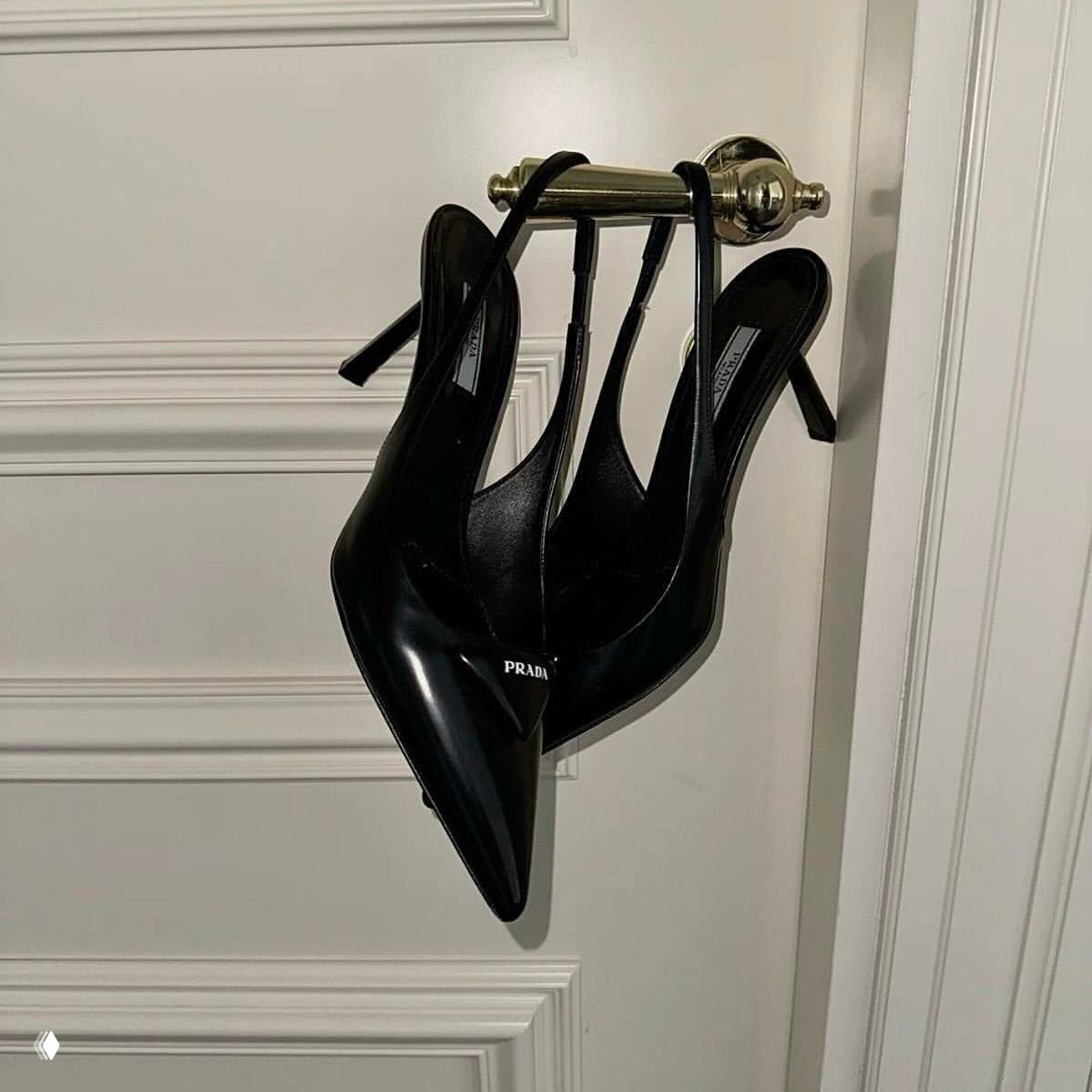 Минималистичный интерьерный кадр: пара чёрных лакированных туфель‑slingback Prada висит на дверной ручке, отражая люксовую, но сдержанную эстетическую деталь.