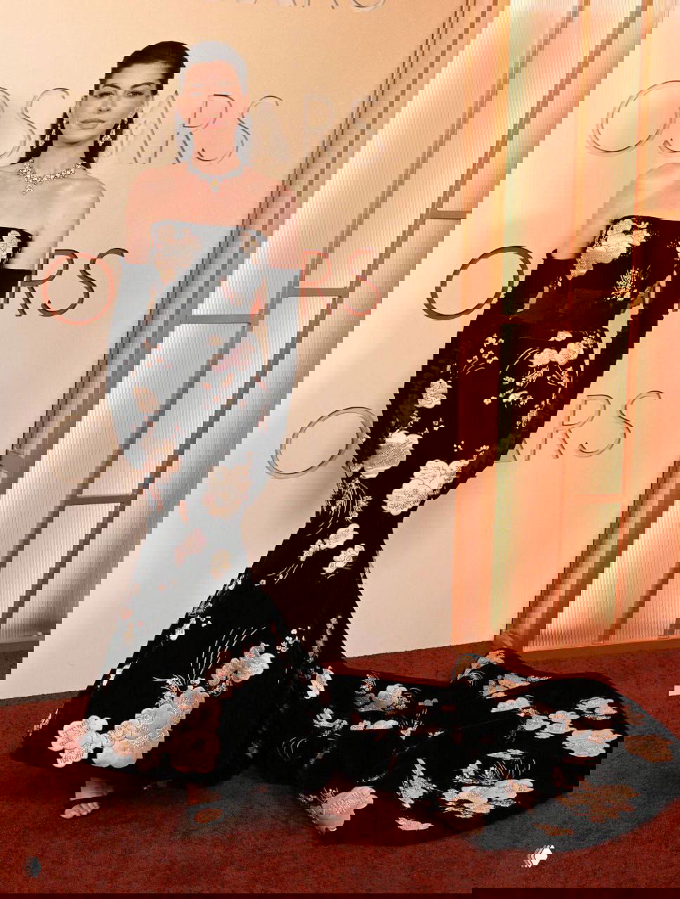 Женщина в чёрном платье «русалка» с цветочным орнаментом и длинным шлейфом, в длинных перчатках, позирует у баннера церемонии.