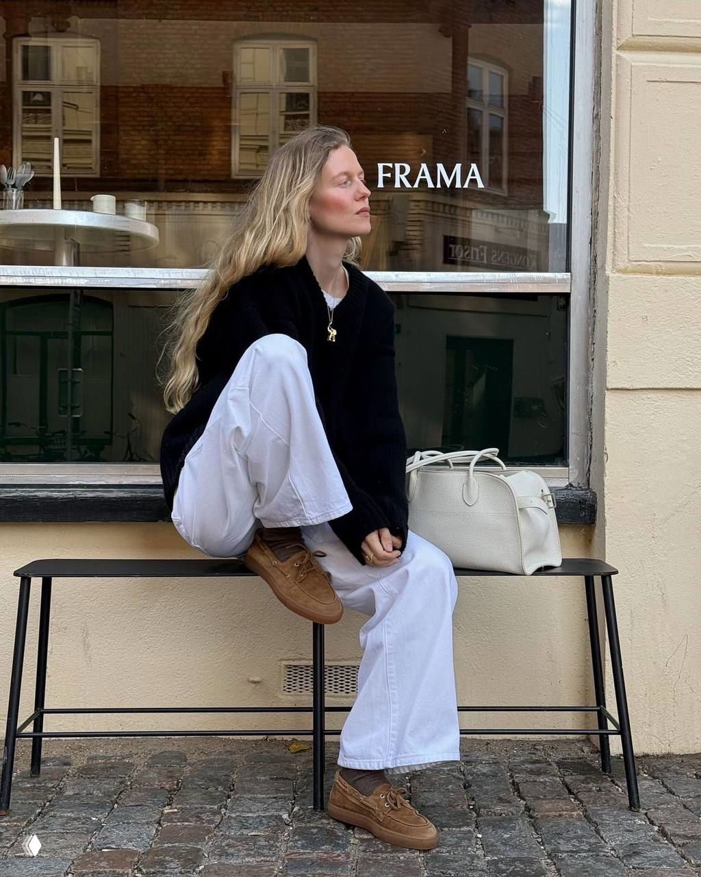 Девушка на скамейке в белых брюках и чёрном свитере демонстрирует коричневые лоферы/кеды — минималистичный скандинавский образ на фоне витрины кафе.