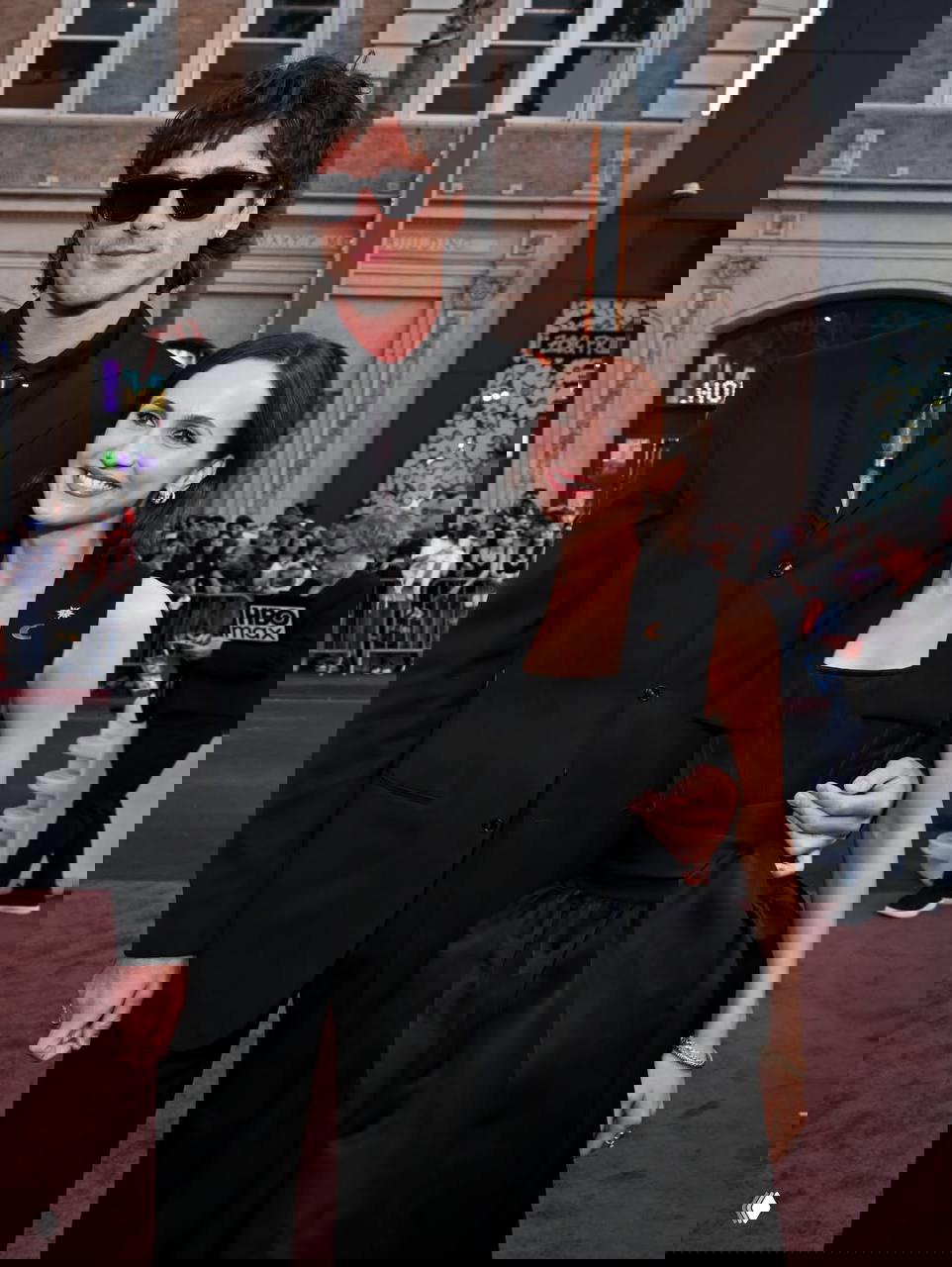 Пара гостей в тёмных нарядах улыбается на красной дорожке премьеры: мужчина в костюме и женщина в вечернем платье среди публики.