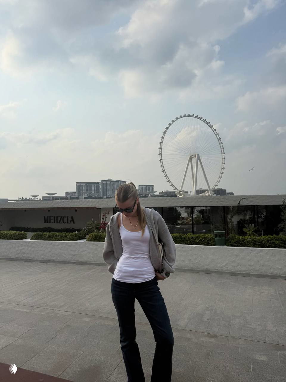 Кристи Крайм стоит на набережной в Дубае на фоне большого колеса обозрения, спокойная поза и городской пейзаж на заднем плане.