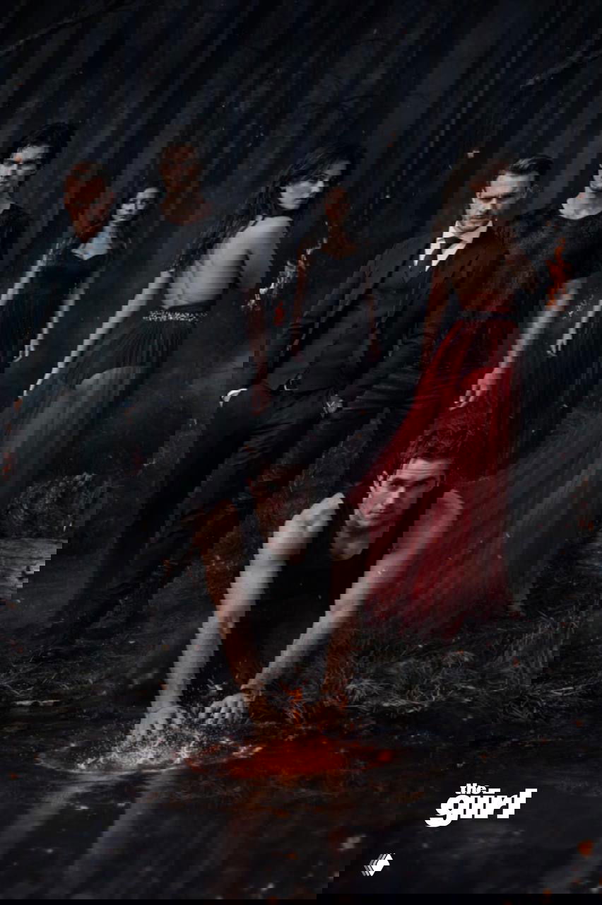 Мрачный лесной кадр: несколько персонажей в тёмной одежде и одна фигура в красном платье, на переднем плане мужчина на коленях у озера — готическая атмосфера.