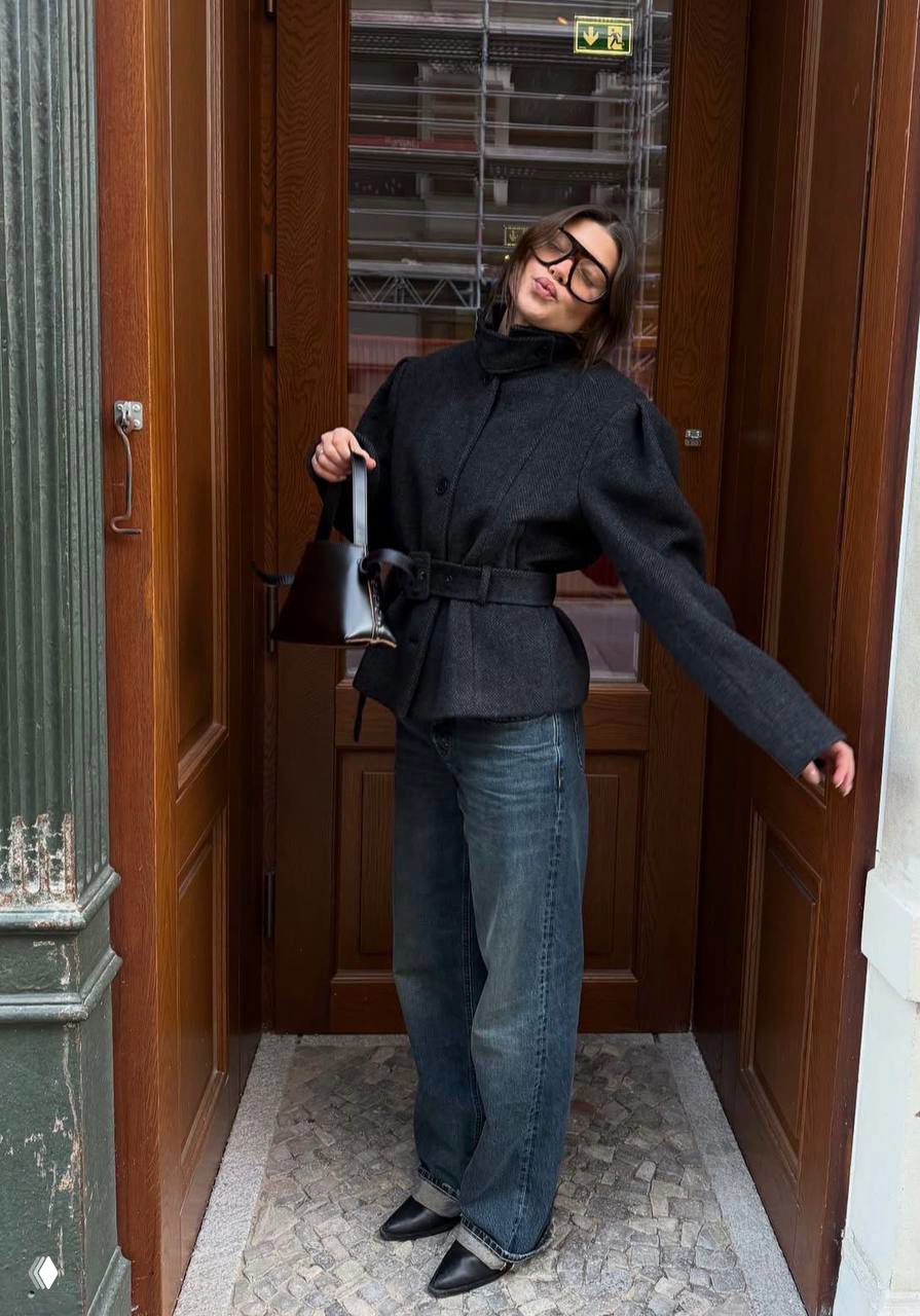 Короткое тёмное приталенное пальто с поясом на фоне деревянной двери; образ с широкими джинсами, сумкой и модными туфлями.
