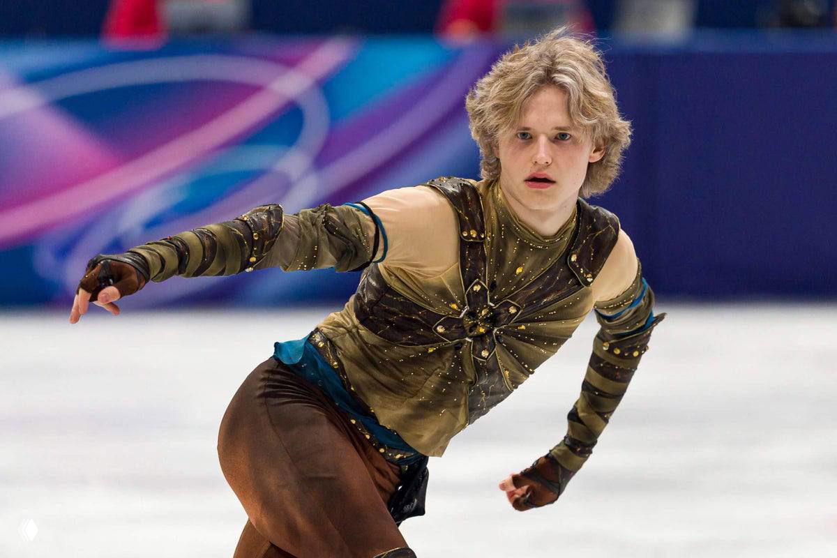 Мужской одиночный фигурист в декоративном костюме выполняет сложный элемент на льду в ярком свете арены и фоне трибун.