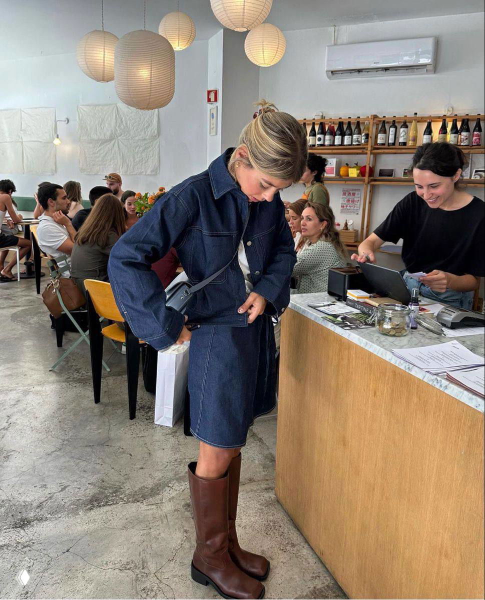 Интерьерное фото кафе: девушка в джинсовом костюме примеряет высокие коричневые сапоги с широким голенищем — стилизация в городской обстановке.