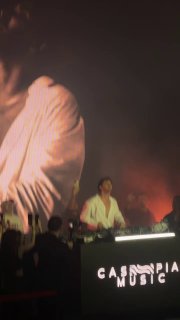 Сцена с DJ Бератом Мертом Демиром в белой рубашке, крупный план диджея, световое шоу и публика перед сценой в клубной атмосфере.