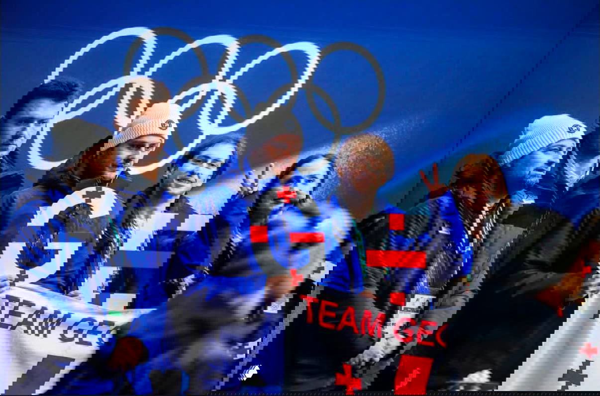 Болельщики с флагами Грузии и баннером TEAM GEO стоят и поддерживают спортсменов у зоны трибун рядом с олимпийскими символами.