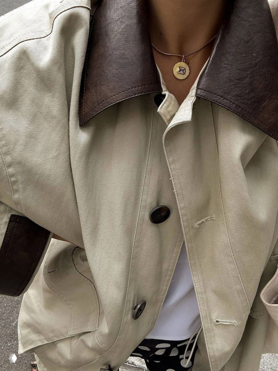 Крупный план бежевой куртки с контрастным коричневым воротником и тонким кулоном на шее; видны фактура ткани и детали отделки.