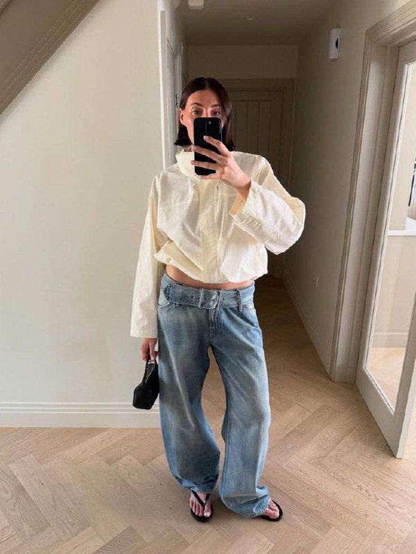 Селфи в светлой укороченной рубашке и широких джинсах, образ с ремнём на талии и минималистичными сандалиями — расслабленный городской стиль.