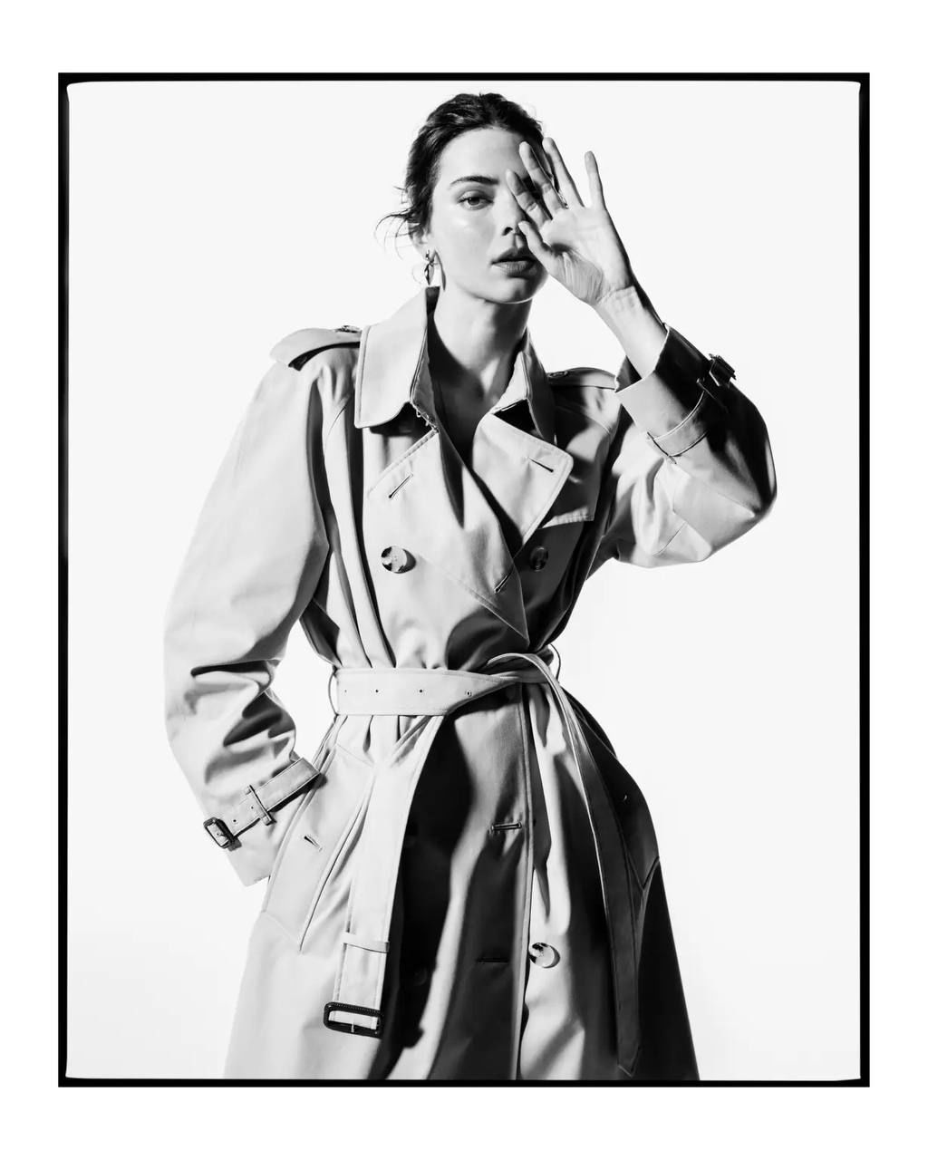 Чёрно‑белый портрет модели в тренчкоте на белом фоне; крупный план, выразительная поза и высокий контраст света и тени.